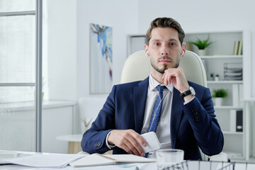 Wall Mural - Portrait of serious business manager in earphones sitting at desk and holding modern phone in office