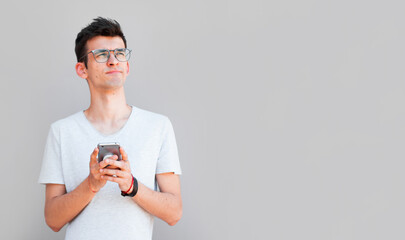 Wall Mural - Portrait of handsome young adult with dreamy look, thinking while holding smartphone, isolated over white background. Son tries to made up message for his father, explaining why he took car