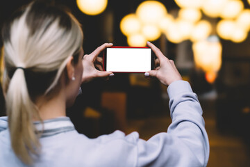 Back view of female holding with hands in horizontal position mobile phone against gold shinning bokeh lights in evening time. Young woman keeps modern smartphone with blank mock up copy space screen