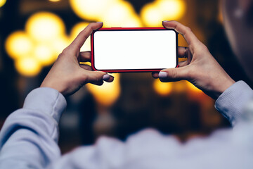 Rear view of female hold in hand in horizontal position mobile phone against gold shinning bokeh lights in evening time. Woman keeps modern smartphone with blank mockup copy space screen for advertise