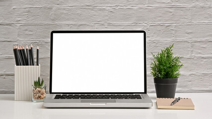 Office creative desk with blank screen laptop, pencil and notebook on white brick wall. Front view