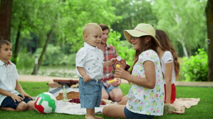 Wall Mural - Parents resting on blanket in park with children. Kids blowing soap bubbles