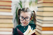 © Roman - Cute smiling preschool age girl in round big glasses with red book in her hands between two books piles