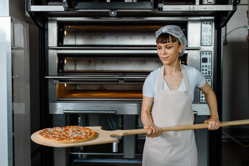 Wall Mural - Young caucasian woman baker is holding a wood peel with fresh pizza near an oven at a baking manufacture factory.