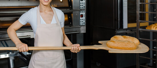 Wall Mural - Close up of young caucasian woman baker is holding a wood peel with fresh bread near an oven at baking manufacture factory.