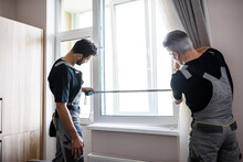 Two Professional Workers In Uniform Using Tape Measure While Measuring Window For Installing Blinds Indoors. Construction And Maintenance Concept