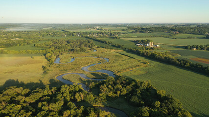 Aerial view of rural landscape with lush greenery, trees, river in the foreground and farmhouse, farmland in the background. American midwestern countryside scenery in the early sunny morning, summer