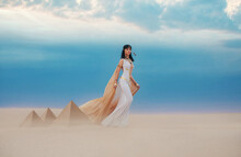  Young Beautiful Woman Model Image Queen Cleopatra Walks In Desert Egyptian Pyramids. Creative Retro Traditional Gold Jewelry Bob Hair Cut. White Satin Long Vintage Dress, Orange Silk Cape Fly Train