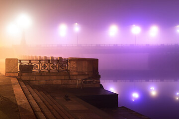 Wall Mural - Illuminated empty Stone bridge in a fog at night. Lanterns close-up. Symmetry reflections on the water, natural mirror. Daugava river, Riga, Latvia. Concept image, neon colors