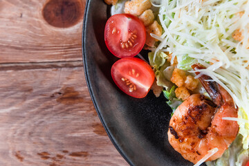 Appetizing Caesar salad with shrimps in a black plate on a wooden table next to a lemon. The concept of healthy food and serving