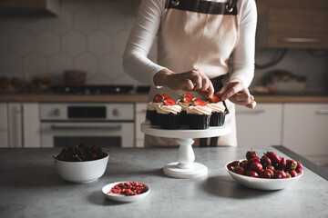 Young woman decorate cupcakes with fruits and cream cheese closeup in kitchen. Working at home. Selective focus.