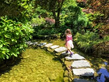 A Young Girl Walking Across Stepping Stones On A Pond In A Beautiful Garden