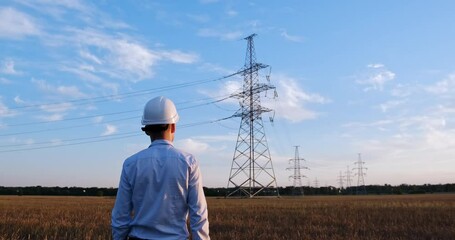 Wall Mural - a man in a white protective helmet goes towards the high-voltage power line poles, stops and looks a