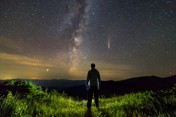 Dark silhouette of a man standing in mountains at night enjoying milky way and Neowise comet with light tail in dark sky view.