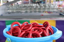 Closeup Of Red Rings In Blue Bucket For Ring Toss At Carnival, With Clear Glass Bottles In Bokeh Effect In Background. Leisure, Fun, Amusement Concepts. Copy Space.