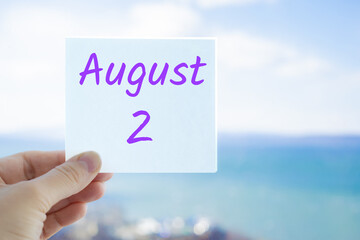 August 2nd. Hand holding sticker with text August 2 on the blurred background of the sea and sky. Copy space for text. Month in calendar concept