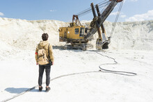 The Traveler Stands In Front Of A Large Excavator. White Sand Pit. Stalker In The World Of Post-apocalypse.