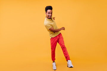 Wall Mural - Full-length portrait of glad african guy in red pants fooling around in studio. Indoor photo of curly black man dancing on yellow background.