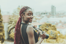 A Dazzling Young African Tourist Woman Photographer With Maroon Colored Braids Is Staying On Some Observation Point, Smiling And Holding A Camera In Her Hands; A Cityscape In The Defocused Background