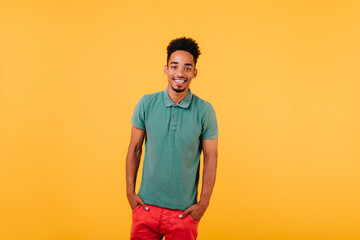 Indoor portrait of smiling black man standing with hands in pockets. Studio shot of blithesome african guy isolated on yellow background.