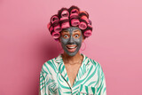 Fototapeta  - Happy joyful woman visits hairdressing and spa salon, makes perfect hairstyle and applies clay face mask, wears pyjama, has surpised expression, isolated on pink background. Female going on date