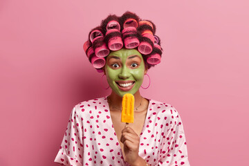 Sticker - Positive woman undergoes beauty procedures, eats yellow popsicle, wears hair curlers for making perfect hairdo, enjoys delicious cold dessert, wears dressing gown, poses against pink background