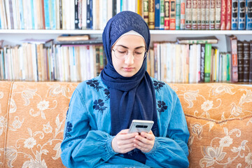 Wall Mural - Happy arabic muslim woman sitting on couch alone using technolog