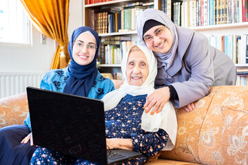 Wall Mural - Happy arabic muslim grandmother and grand daughter and mother sitting togther on couch using technology