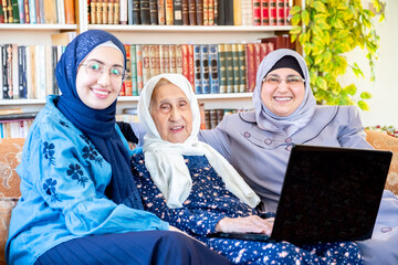 Happy arabic muslim grandmother and grand daughter and mother sitting togther on couch using technology