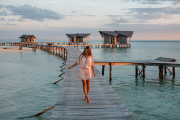 Wall Mural - Young woman in white clothes walking on a pier on Maldives, sunset time