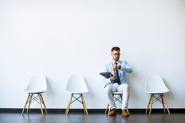Wall Mural - Young man sitting in the waiting room with a folder in hand and checking time before an interview
