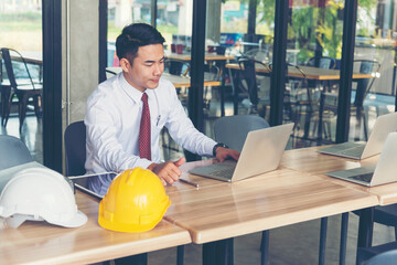 Civil construction engineer working with laptop at desk office with white yellow safety hard hat at office on construction site. Asian young man architecture project manager sitting at office on site