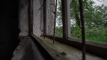 Close Motion Along Broken Window With Rusty Metal Bars Of Abandoned Building Against Waving Trees Outside Slow Motion