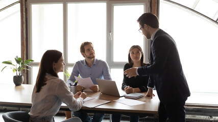 Wall Mural - Motivated diverse colleagues gather at desk in boardroom talk brainstorm considering paperwork together, multiracial employees cooperate work in group on laptop, discuss project, teamwork concept