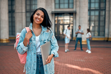 Wall Mural - Cute African American girl student with a backpack and laptop near the campus against the background of a group of students.