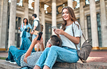 Wall Mural - A couple of students with backpacks and a laptop sit on the steps outside the campus and use their smartphones.