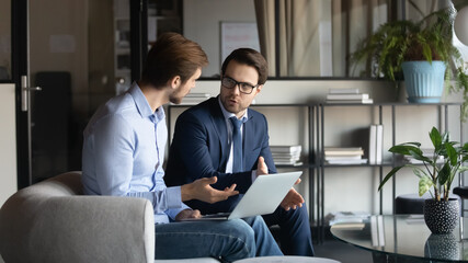 Canvas Print - Concentrated Caucasian male employees sit on couch in office brainstorm work together on laptop, focused businessmen or colleagues talk discuss business ideas cooperating on computer at workplace