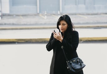 Wall Mural - Young female wearing a black jacket fixing her makeup on the sidewalk of an urban area