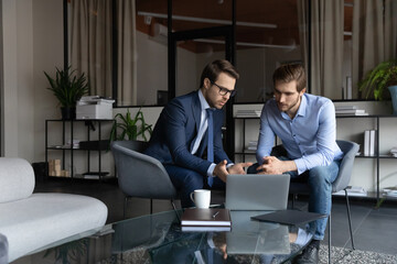 Wall Mural - Serious young caucasian businessmen look at laptop screen brainstorm discuss company financial project in office together, focused male colleagues or business partners cooperate work on computer