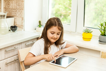 Wall Mural - Little girl at home in the kitchen sitting at the table with tablet