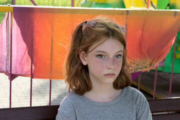 A beautiful teenage girl with ponytails and freckles flushed from the heat. Red-haired girl with a pensive look is sad alone, sitting on a bench in an amusement park in summer.