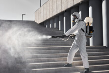 Cleaning, Disinfection Of The City. Professional Worker Man In White Protective Suit Cleans The Stairs With Spray, Sanitation And Disinfection Service