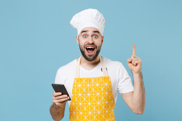 Wall Mural - Excited young bearded male chef or cook baker man in apron white t-shirt toque chefs hat isolated on blue background. Cooking food concept. Using mobile phone hold index finger up with great new idea.