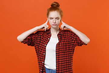 Displeased young readhead girl in casual red checkered shirt posing isolated on orange background studio. People lifestyle concept. Mock up copy space. Covering ears with fingers, keeping eyes closed.