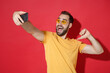 © ViDi Studio - Excited young bearded man guy in casual yellow t-shirt glasses isolated on red background. People lifestyle concept. Mock up copy space. Doing selfie shot on mobile phone pointing thumb on himself.