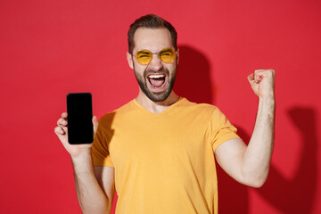 Sticker - Screaming young bearded guy in casual yellow t-shirt glasses isolated on red background. People lifestyle concept. Mock up copy space. Hold mobile phone with blank empty screen doing winner gesture.