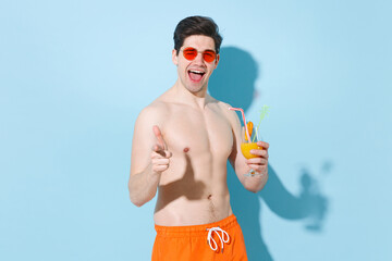 Blinking young man guy in glasses isolated on blue background studio portrait. People summer vacation rest lifestyle concept. Mock up copy space. Hold glass with cocktail point index finger on camera.