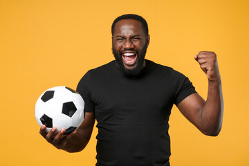 Wall Mural - Screaming african american man guy football fan in black t-shirt isolated on yellow background. Sport family leisure lifestyle concept. Cheer up support favorite team with soccer ball, clenching fist.