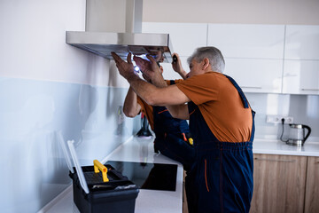 Two workers, handyman in uniform installing or repairing a kitchen extractor, replacing filter in cooker hood. Construction, maintenance and repair concept