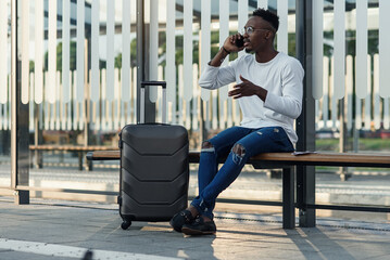 Young handsome Caucasian man in hat with backpack talking with smile on cellphone. Smiling cheerfully at train station. Attractive male traveller speaking on mobile phone at bus stop. Telephone call.